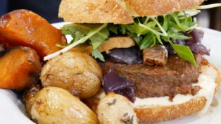 Zoom sur un burger. On voit son accompagnement (patates et patate douce). À l'intérieur du burger, un steak Beyond Meat, des morceaux de figues, de faux gras, et de la roquette.
