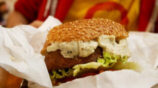 Un burger avec du pain au sésame, de la crème, un épais steak Beyond Meat et de la salade. Au fonc, flou, on voit Guillaume dans son sweat rouge et son t-shirt Noam Chomsky jaune et rouge.