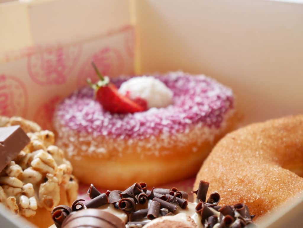 Donuts dans une boite : au fond, flou, le donut Framboise. Au 1er plan, net des copeaux de chocolat sur le donut Kinder Bueno.